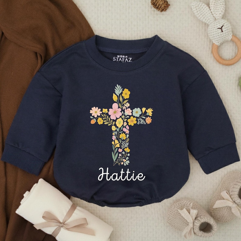 Op de afbeelding: Marineblauwe babyromper met een bloemenkruisontwerp. Het kruis bestaat uit kleurrijke bloemen in roze, geel en blauw. De naam "Hattie" staat in sierlijke letters eronder. De romper heeft lange mouwen en een ronde halslijn.