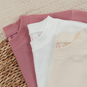 Op de afbeelding: Drie crewneck sweatshirts in roze, witte en cr&egrave;metinten. De sweatshirts zijn opgevouwen, met de kragen zichtbaar. De tekst "FA" is gedrukt op het cr&egrave;mekleurige sweatshirt. De stof lijkt zacht en comfortabel.