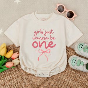 May include: A white onesie with short sleeves and a pink bow. The onesie has the text "girls just wanna be one" printed on it.
