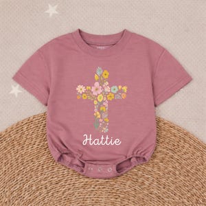 Op de afbeelding: Stofroze babyromper met een bloemenkruisontwerp. Het kruis bestaat uit diverse kleurrijke bloemen. De naam "Hattie" staat in cursieve letters eronder. De romper heeft korte mouwen en drukkers aan de onderkant.