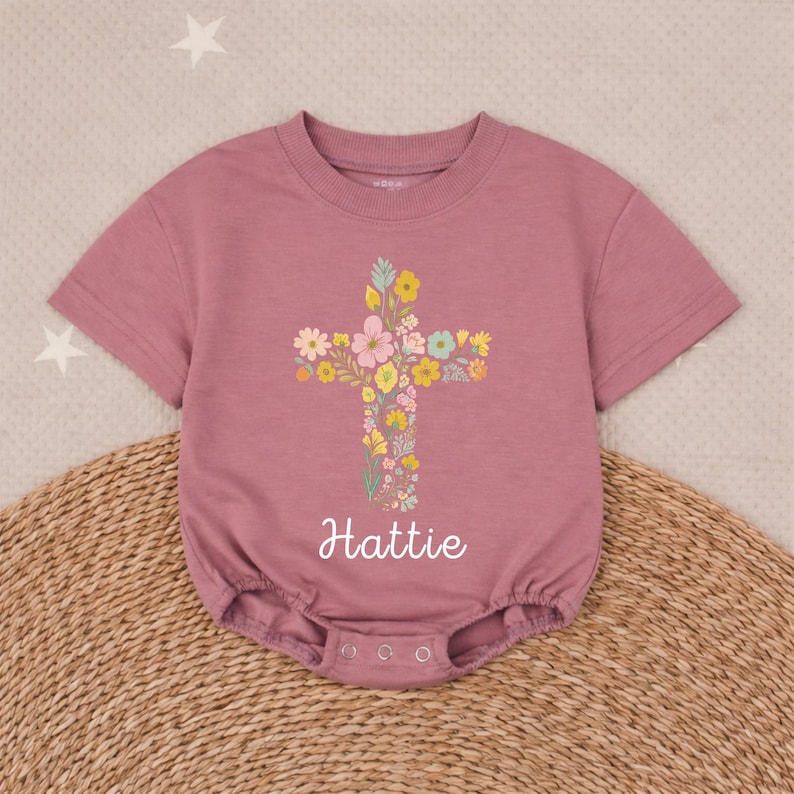 Op de afbeelding: Stofroze babyromper met een bloemenkruisontwerp. Het kruis bestaat uit diverse kleurrijke bloemen. De naam "Hattie" staat in cursieve letters eronder. De romper heeft korte mouwen en drukkers aan de onderkant.