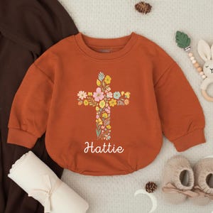 Op de afbeelding: Roestkleurige babyromper met een bloemenkruisontwerp. Het kruis is gevuld met kleurrijke bloemen en bladeren. De naam "Hattie" staat eronder. De romper wordt getoond met babyschoentjes en een opgerolde deken.