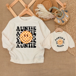 May include: A white sweatshirt with the text "AUNTIE" repeated in a retro font with a checkered smiley face in the center. A smaller white sweatshirt with the text "AUNTIE'S BESTIE" and a checkered smiley face is also shown.