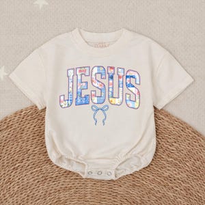 May include: Cream-colored baby onesie with the word "JESUS" in a patchwork design of blue, pink, and yellow floral and gingham patterns. A blue bow is below the word. The onesie has short sleeves and snaps at the bottom.
