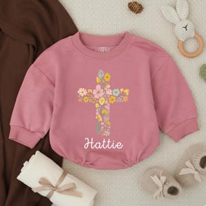 Op de afbeelding: Een oudroze babyromper met een bloemenkruisontwerp. Het kruis is gemaakt van kleurrijke bloemen. De naam "Hattie" staat eronder gedrukt. Babyaccessoires omringen de romper.