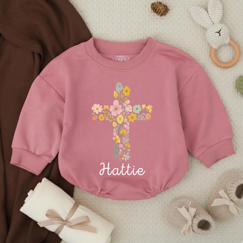 Op de afbeelding: Een oudroze babyromper met een bloemenkruisontwerp. Het kruis is gemaakt van kleurrijke bloemen. De naam "Hattie" staat eronder gedrukt. Babyaccessoires omringen de romper.