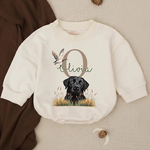 May include: Cream-colored baby romper with a graphic of a black Labrador Retriever, a flying duck, and the name "Olivia" in script. The romper has long sleeves and a round neckline. The brand name "Mosa Tafaz" is printed on the neckline.