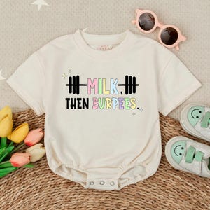 May include: Cream-colored baby onesie with the text "MILK, THEN BURPEES." in colorful letters and a barbell graphic. The onesie has short sleeves and snaps at the bottom. Pink sunglasses and baby shoes are also in the image.