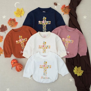 Op de afbeelding: Een collectie kindersweatshirts in verschillende kleuren: marineblauw, roest, cr&egrave;me en mauve. Elk sweatshirt heeft een bloemenkruisontwerp op de voorkant. De naam "Hattie" staat op het witte sweatshirt. Herfstbladeren en decoratieve elementen omringen de sweatshirts.