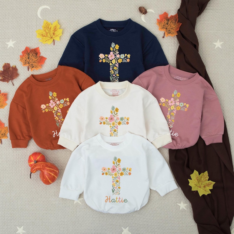 Op de afbeelding: Een collectie kindersweatshirts in verschillende kleuren: marineblauw, roest, cr&egrave;me en mauve. Elk sweatshirt heeft een bloemenkruisontwerp op de voorkant. De naam "Hattie" staat op het witte sweatshirt. Herfstbladeren en decoratieve elementen omringen de sweatshirts.