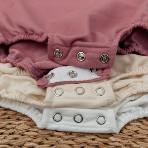 Op de afbeelding: Close-up van verschillende babyrompers in tinten roze, cr&egrave;me en wit. De rompers hebben drukknoopsluitingen en elastische taillebanden. De stof lijkt zacht en comfortabel, geschikt voor een baby.