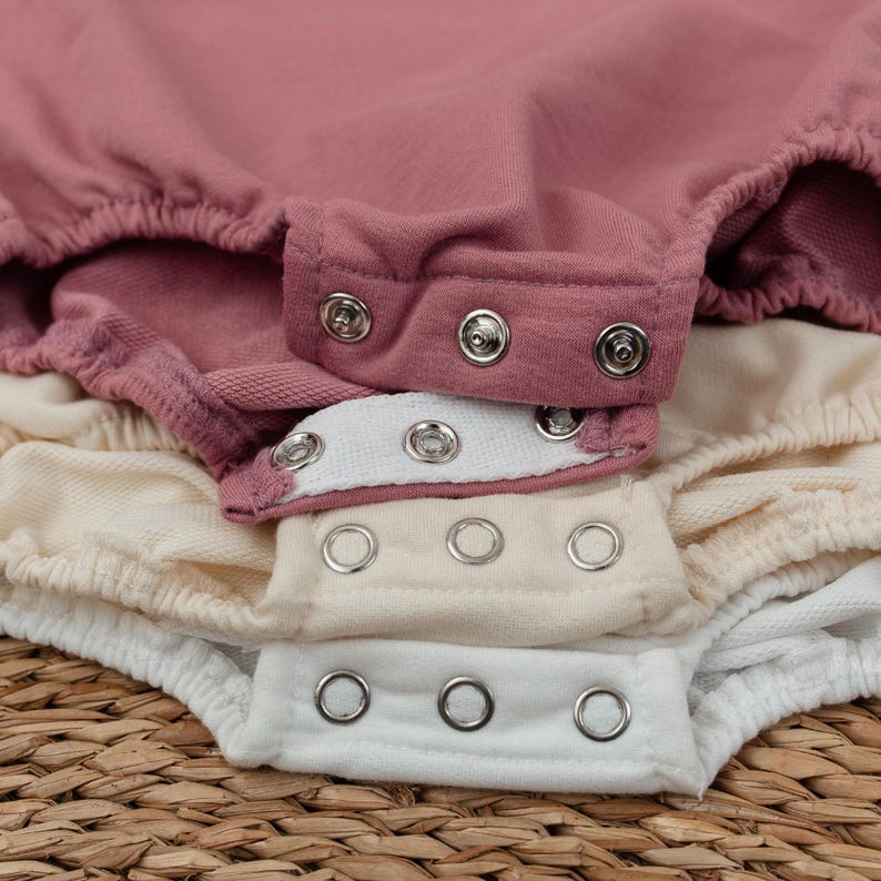 Op de afbeelding: Close-up van verschillende babyrompers in tinten roze, cr&egrave;me en wit. De rompers hebben drukknoopsluitingen en elastische taillebanden. De stof lijkt zacht en comfortabel, geschikt voor een baby.