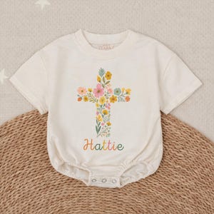Op de afbeelding: Cr&egrave;mekleurig babyrompertje met een bloemenkruisontwerp. Het kruis bestaat uit kleurrijke bloemen en groen. De naam "Hattie" staat in een regenbooglettertype eronder. Het rompertje heeft korte mouwen en drukkers aan de onderkant.