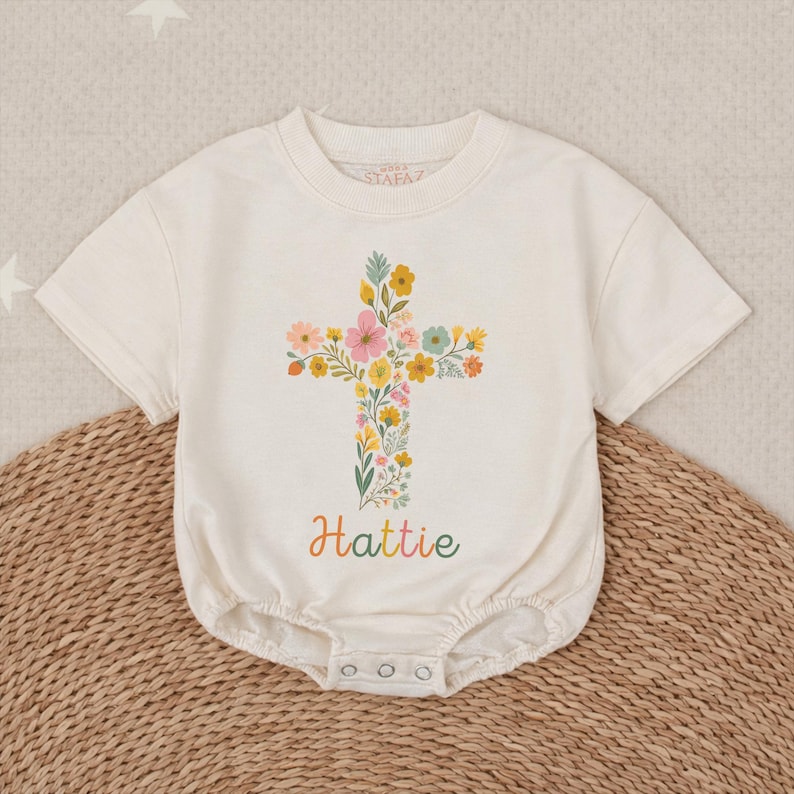 Op de afbeelding: Cr&egrave;mekleurig babyrompertje met een bloemenkruisontwerp. Het kruis bestaat uit kleurrijke bloemen en groen. De naam "Hattie" staat in een regenbooglettertype eronder. Het rompertje heeft korte mouwen en drukkers aan de onderkant.