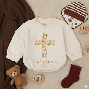 Op de afbeelding: Cr&egrave;mekleurige babyromper met een bloemenkruisontwerp en de naam "Hattie" in kleurrijke letters. De romper wordt gecombineerd met een bruin-wit gestreepte muts, een slab, een paar bordeauxrode sokken en een teddybeer.