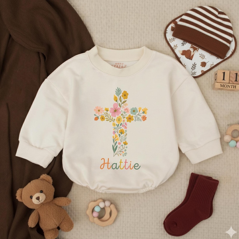 Op de afbeelding: Cr&egrave;mekleurige babyromper met een bloemenkruisontwerp en de naam "Hattie" in kleurrijke letters. De romper wordt gecombineerd met een bruin-wit gestreepte muts, een slab, een paar bordeauxrode sokken en een teddybeer.