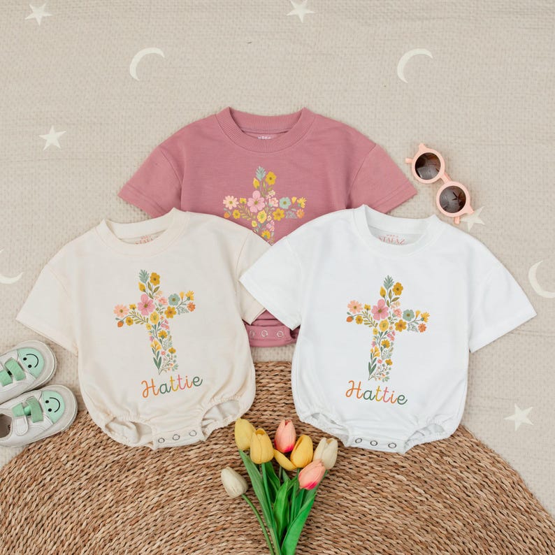 Op de afbeelding: Drie baby-outfits: een roze shirt met korte mouwen en twee rompers met korte mouwen in cr&egrave;me en wit. Elk heeft een bloemenkruisontwerp en de naam "Hattie" in kleurrijk schrift. Zonnebril en schoenen zijn ook zichtbaar.