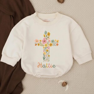 Op de afbeelding: Cr&egrave;me babyromper met een bloemenkruisontwerp. Het kruis is gevuld met kleurrijke bloemen en groen. De naam "Hattie" staat in een regenbooglettertype eronder. De romper heeft lange mouwen en een ronde halslijn.