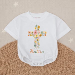 Op de afbeelding: Witte babybody met een bloemenkruisontwerp. Het kruis bestaat uit diverse kleurrijke bloemen en groen. De naam "Hattie" staat in een kleurrijk handschrift eronder. De body heeft korte mouwen en drukknoopsluitingen.
