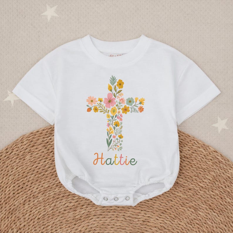 Op de afbeelding: Witte babybody met een bloemenkruisontwerp. Het kruis bestaat uit diverse kleurrijke bloemen en groen. De naam "Hattie" staat in een kleurrijk handschrift eronder. De body heeft korte mouwen en drukknoopsluitingen.