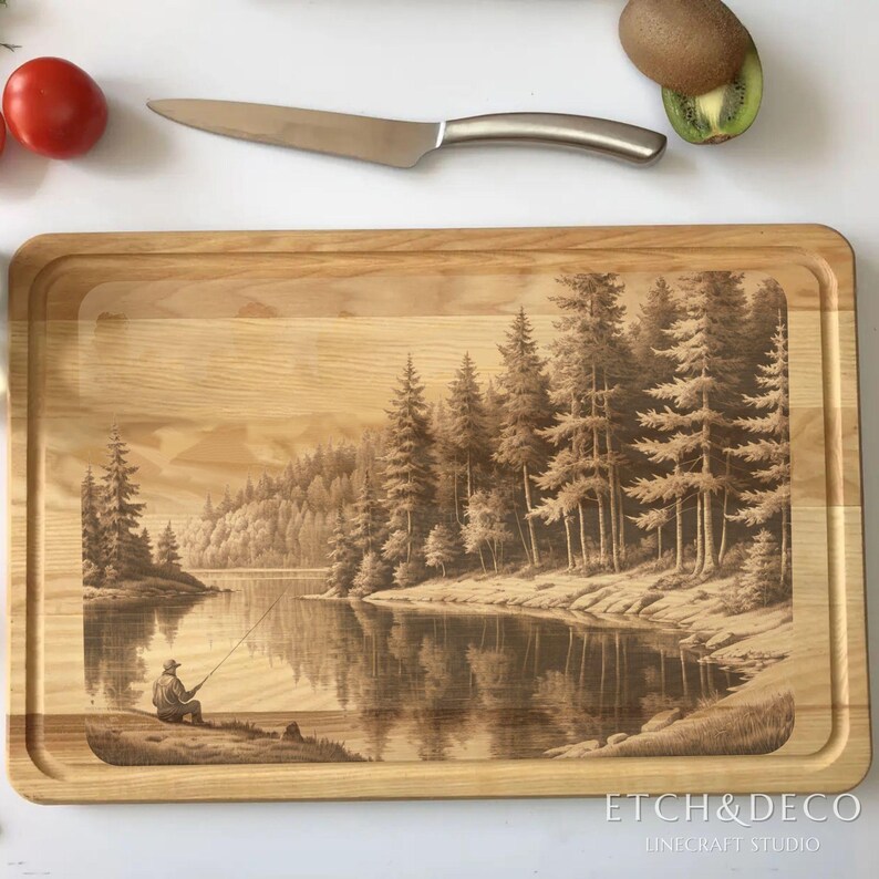 May include: A rectangular wooden cutting board with a detailed engraved scene of a person fishing on a lake surrounded by trees. A stainless steel knife and a tomato are visible. The words "ETCH&DECO" and "LINECRAFT STUDIO" are at the bottom.