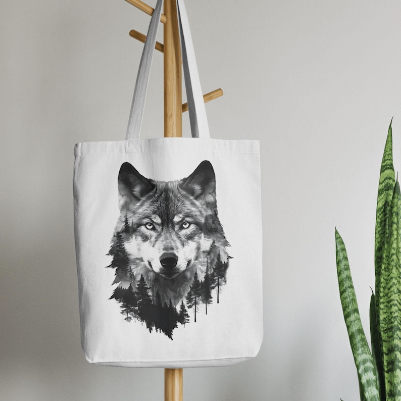 May include: White tote bag featuring a detailed black and white illustration of a wolf's face, with a forest scene integrated into the design. The bag has a simple, minimalist design with long handles.
