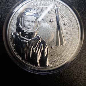 May include: A circular silver commemorative coin encased in a clear plastic capsule. The coin features a relief portrait of Yuri Gagarin in a space suit, a rocket, a globe, and stars, with Cyrillic text inscribed along the upper and right edges.