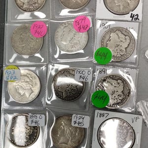May include: A display of vintage silver coins, each encased in a protective sleeve. The coins vary in date and design, with some featuring profiles and inscriptions. Price tags in various colors are attached to the sleeves.