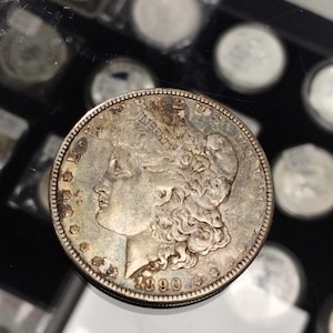 May include: A silver Morgan dollar coin from 1890, featuring a profile of Lady Liberty and the inscription "Liberty." The coin has a detailed design with visible wear and tear, and is surrounded by other coins.