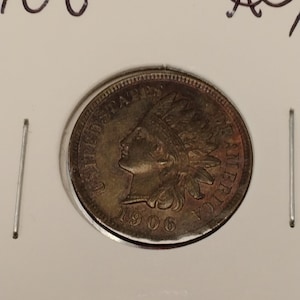 May include: A 1906 Indian Head penny, displayed in a protective holder. The coin features a profile of a Native American in a feathered headdress and the words "UNITED STATES OF AMERICA". The year "1906" is visible on the coin.