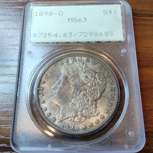 May include: A silver 1898-O Morgan silver dollar coin, encapsulated in a clear plastic holder. The coin features a profile of a woman and the text "E PLURIBUS UNUM". The holder has a label with the text "MS63" and "S $1".
