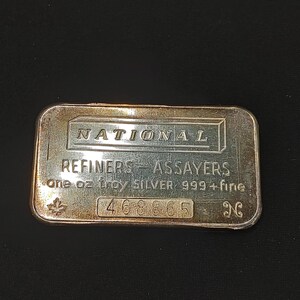 May include: A rectangular silver bullion bar with a metallic finish. The text reads NATIONAL REFINERS-ASSAYERS, one oz troy SILVER 999+ fine, and the serial number 468865. A small maple leaf symbol and an N logo are embossed on the front surface.