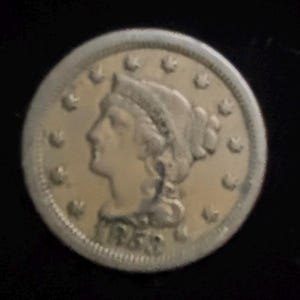 May include: A silver coin with a profile of a woman wearing a head covering, surrounded by stars. The coin has the year 1853 inscribed on it. The coin is set against a black background.