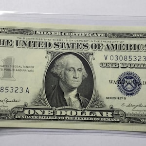 May include: A one dollar silver certificate from 1957. The certificate features a portrait of George Washington and the text "The United States of America" in bold lettering. The serial number is V 03085323 A.