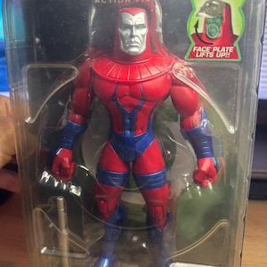 May include: A Green Lantern action figure in a clear plastic package. The figure is primarily red and blue, with a silver face and a helmet. The packaging includes the text "Green Lantern" and "Action Figures."