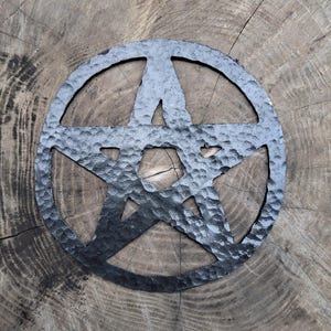 Pode incluir: Um pentagrama de parede circular em metal preto com um acabamento martelado e texturizado. A estrela de cinco pontas está inserida numa moldura circular sólida, exibindo uma aparência metálica escura com reentrâncias visíveis.