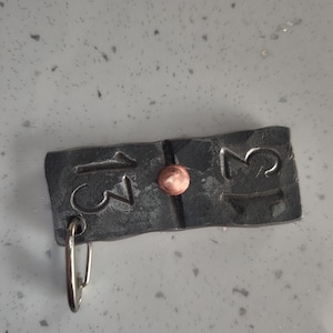 May include: Hand-forged metal keyring with the number "13" on each side of a copper rivet. The dark grey metal has a hammered texture and a silver-coloured key ring attached to one end. A unique, handcrafted accessory.