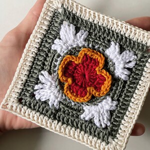 May include: A square crochet piece featuring a central red flower with orange trim, surrounded by white petal-like accents. The design is set against a green background, framed by a cream-colored border. The handmade item showcases a floral theme.
