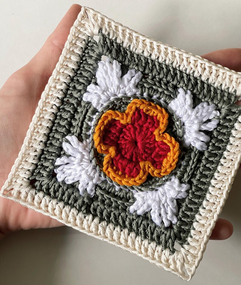 May include: A square crochet piece featuring a central red flower with orange trim, surrounded by white petal-like accents. The design is set against a green background, framed by a cream-colored border. The handmade item showcases a floral theme.
