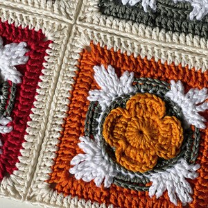 May include: Close-up of a crocheted blanket composed of square motifs. Each square features a central flower design in orange, surrounded by white, green, and cream-colored yarn. The squares are bordered in red, orange, and cream.