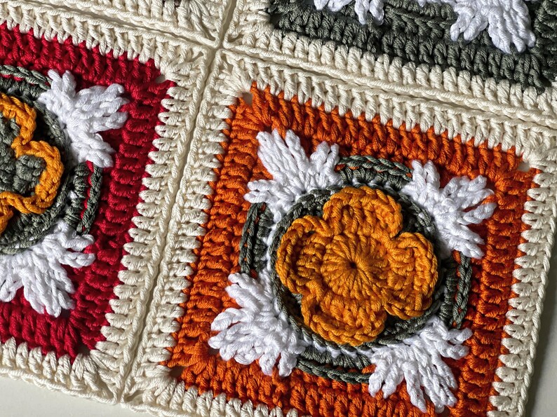 May include: Close-up of a crocheted blanket composed of square motifs. Each square features a central flower design in orange, surrounded by white, green, and cream-colored yarn. The squares are bordered in red, orange, and cream.