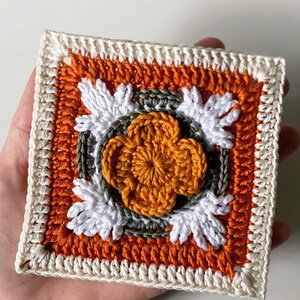 May include: A square, handmade crochet piece featuring a central orange flower design surrounded by white and green accents. The square has an orange background and a cream-colored border. The piece is held in a hand.