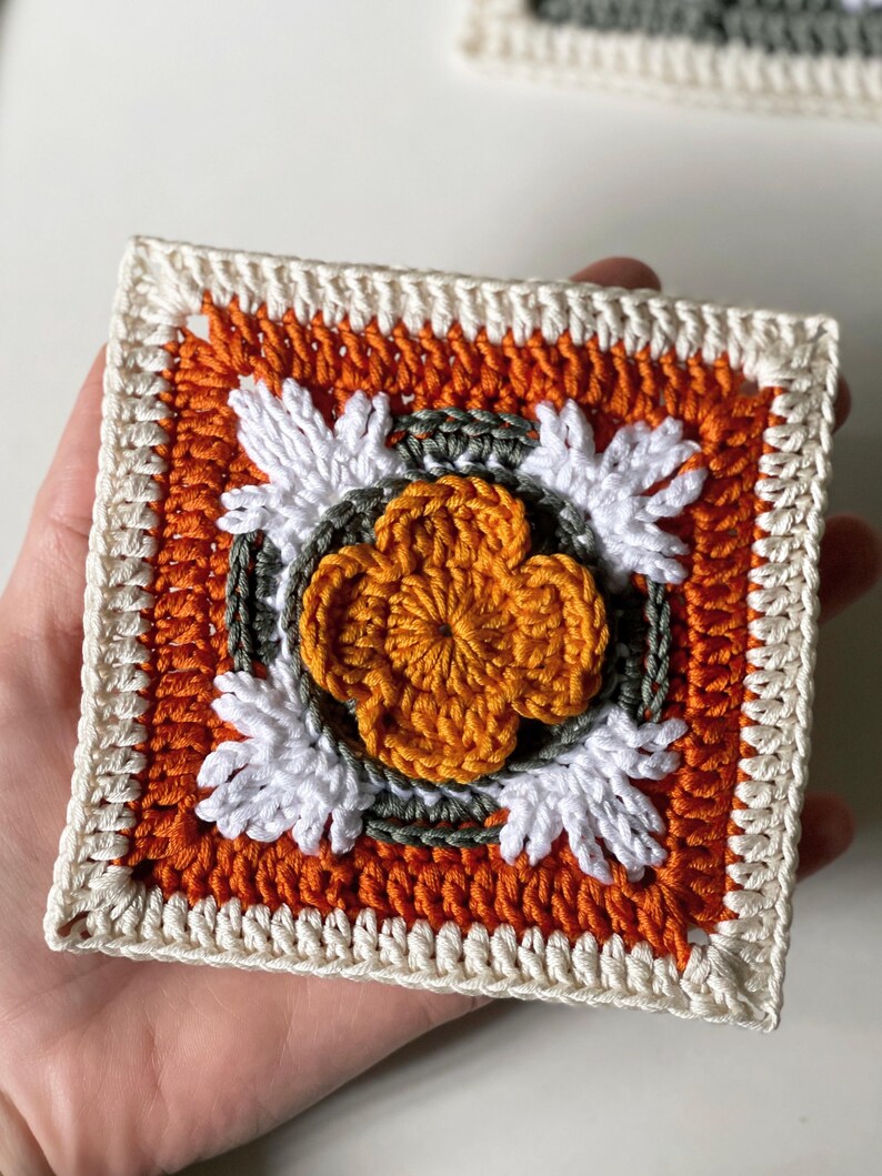 May include: A square, handmade crochet piece featuring a central orange flower design surrounded by white and green accents. The square has an orange background and a cream-colored border. The piece is held in a hand.