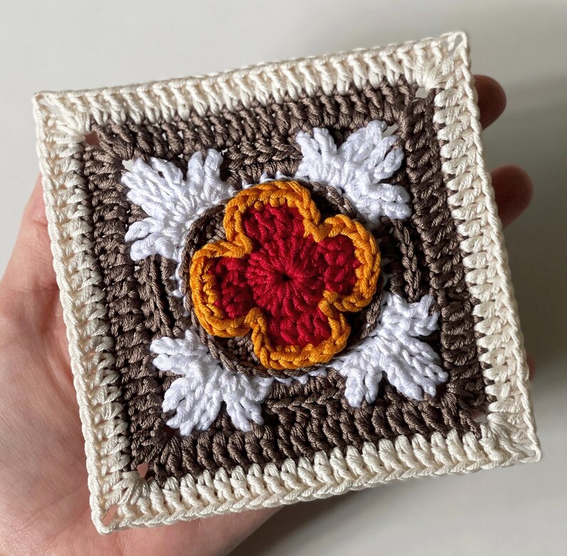 May include: A square, handmade crochet piece featuring a central red and orange flower design. The flower is surrounded by white leaf-like shapes, set against a brown background, and framed by a cream-colored border.