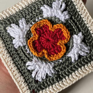 May include: A square, handmade crochet piece featuring a central red flower with orange trim, surrounded by white and green floral accents. The border is cream-colored. This item is a handcrafted textile, perfect for home decor or crafting projects.