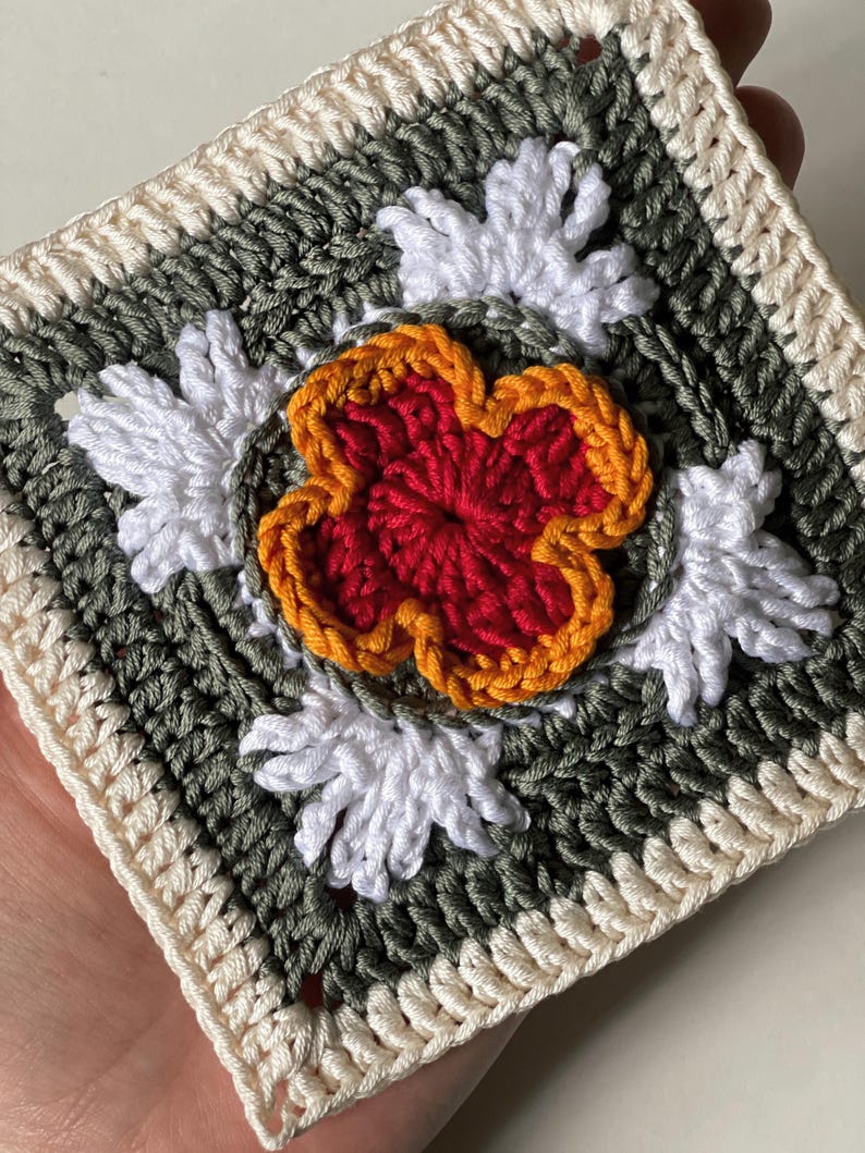 May include: A square, handmade crochet piece featuring a central red flower with orange trim, surrounded by white and green floral accents. The border is cream-colored. This item is a handcrafted textile, perfect for home decor or crafting projects.