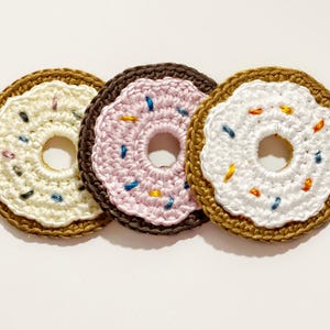 Könnte beinhalten: Drei handgefertigte Häkel-Donut-Untersetzer. Jeder Untersetzer hat ein anderes Farbschema: Vanille mit Streuseln, Rosa mit Streuseln und Weiß mit Streuseln. Die Untersetzer haben einen braunen Rand und ein Loch in der Mitte.