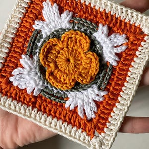 May include: A square crochet piece featuring a central orange flower with white petal accents. The design includes orange, white, and green yarn, with a cream-colored border. The handmade item showcases a detailed floral pattern.