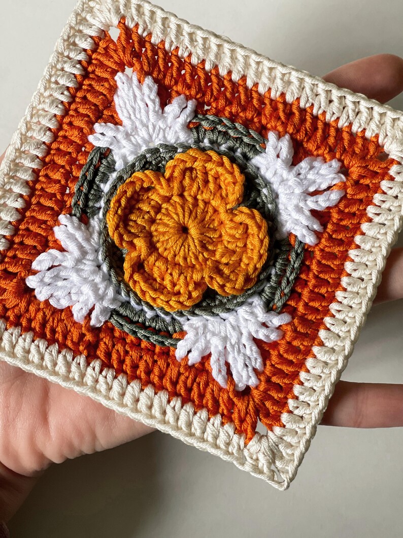 May include: A square crochet piece featuring a central orange flower with white petal accents. The design includes orange, white, and green yarn, with a cream-colored border. The handmade item showcases a detailed floral pattern.