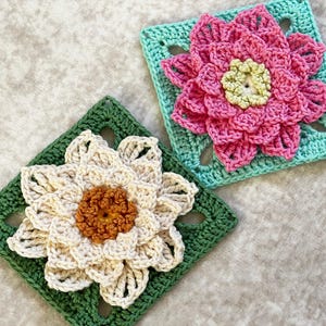 May include: Two handmade crochet flower squares. One features a pink flower with a yellow center on a turquoise square. The other has a cream-colored flower with a brown center on a green square. The squares are on a light gray surface.